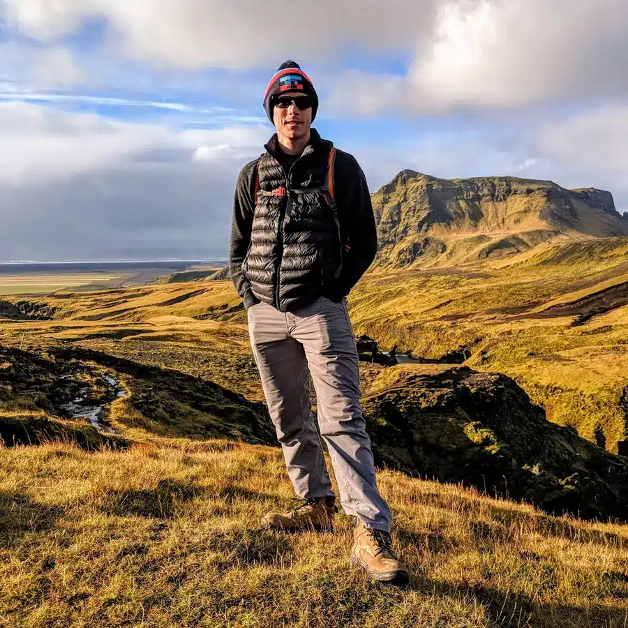 Brett Folkerts standing on a grassy hillside in Iceland in a beanie, sunglasses and down vest, with green mountains behind him.