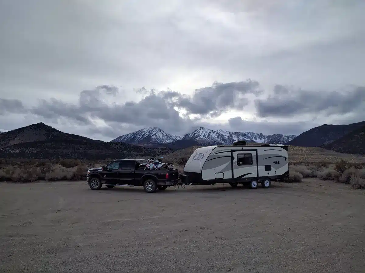 Forest River Vibe travel trailer hitched to a black Ford F-250 pickup carrying a dirt bike, parked on a gravel flat below snow-capped mountains.
