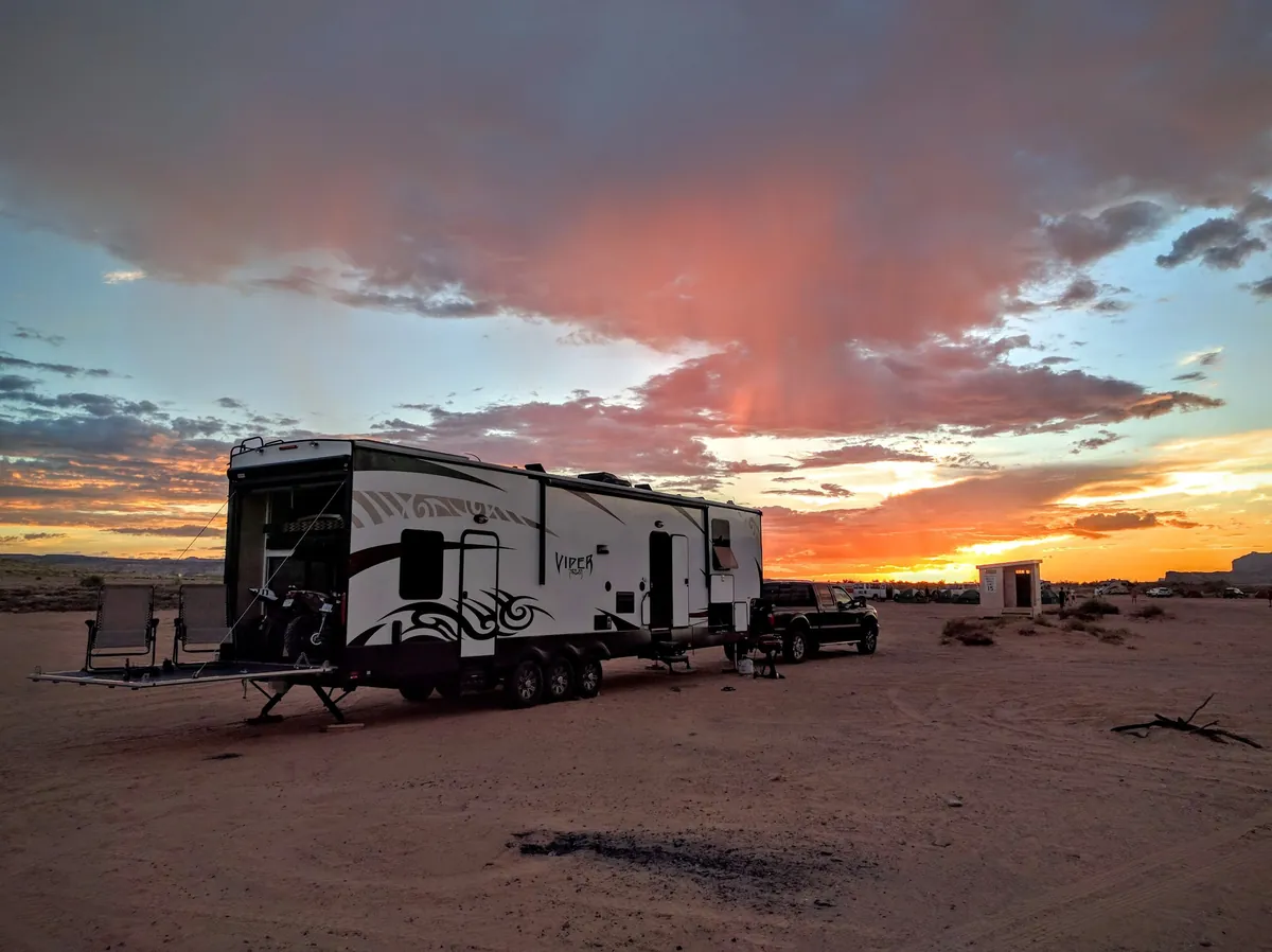 Forest River toy hauler fifth wheel parked in the desert at sunset with its rear ramp lowered, chairs on the ramp and motorcycles inside.