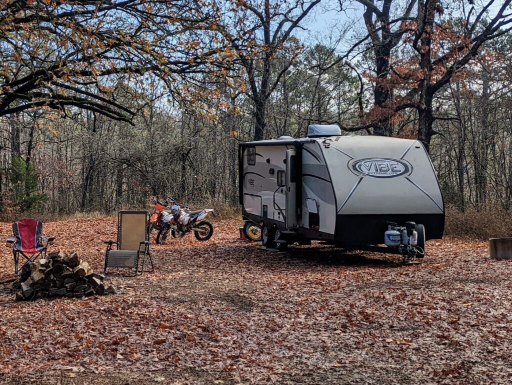 Vibe travel trailer parked under fall trees with two dirt bikes, camp chairs and a stack of firewood